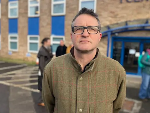 Tom Rainey in front of The King's Centre on Osney Mead. He is wearing glasses. There are other people behind him