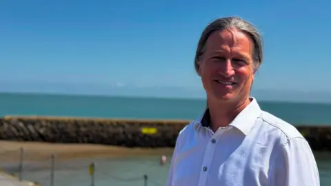 A smiling man with tied-back long grey hair wearing a white shirt. He is standing next to the sea.