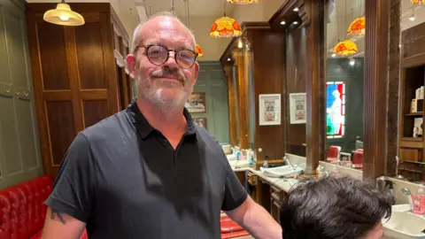 Simon Thake/BBC A barber looking into the camera while a customer on the chair in front of him stares at the mirror straight ahead. The barber has glasses, a short beard and a black polo shirt. The barbershop behind him is brightly lit and has oak furnishings. 