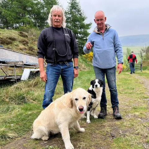 Two men - one wearing a dark blue top and jeans, the other light blue top and jeans, stand behind a golden retriever and a Border collie, on a dirt track with a woodland behind them