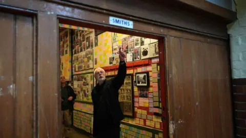 Getty Images A white-bearded man in an all-black outfit points up inside a room covered with photographs. The Photo is taken through the wood panels of an open door, with a sign above the opening reading "Smiths"