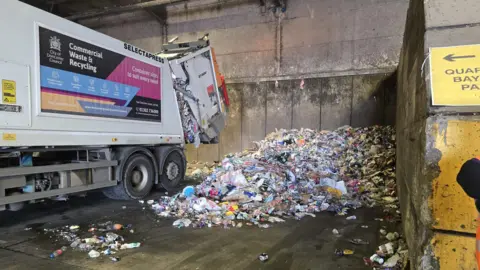 Hayley Coyle/BBC A bin lorry at the depot with its back partially opened to allow rubbish to fall out. Behind the truck is a large pile of refuse of all different colours and sizes.