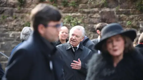 Athena A man in a black jacket and tie and white hair is standing speaking 