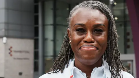 Ijeoma Okolo wears a white shirt with a lanyard around her neck. She has braided hair and is smiling at the camera. She stands in front of the Gateway building at the University of Essex's Southend campus.