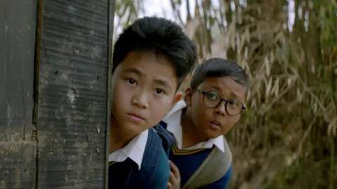Excel Entertainment, Chalkboard Entertainment & Suitable Pictures Two boys in school uniforms - one behind the other - looking at something, partially taking cover of a wooden surface. The one behind wears spectacles and is carrying a school bag.