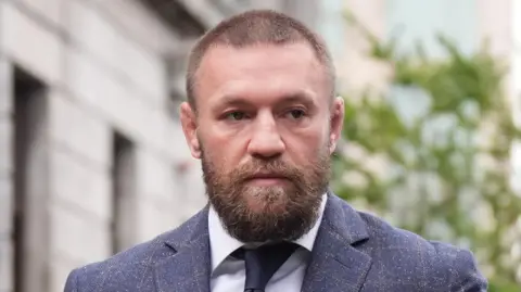 Conor McGregor outside court on Tuesday, he is wearing a navy tweed suit jacket with a white shirt and navy tie. He is short brown hair and a brown beard. 