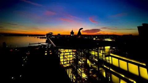 A beautiful sunset over the Senedd building in Cardiff Bay. The photograph is over the top of the building from the back, looking out across the bay. The skyline is a mixture of blue, orange and red and the yellow lights in the building contrast strongly with the darkness immediately outside the building.