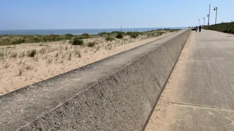 A large concrete wall runs along a beach. There is a footpath on one side and sand dunes planted with grasses on the other side. The sea can be seen under a blue sky.
