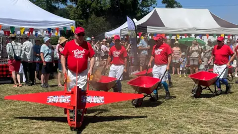 Barlow Red Barrows Four people in red t shirts pushing red wheelbarrows in formation. A crowd looks on in the background and two white marquee roofs can be seen.