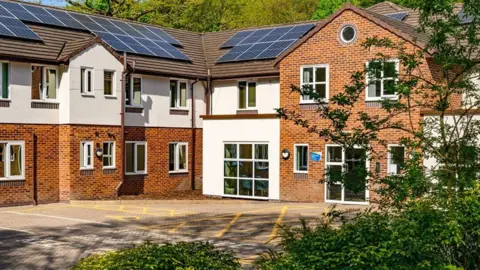 Cygnet Hospital Kidsgrove has two storeys. In the photo, part of the building is at right angles to another section of the building. Trees are behind it and one tree is in front of the facility on the right hand side.