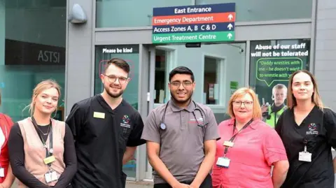 Ali Sidat stands outside a hospital entrance with a stethoscope around his neck, in the middle of a group of five colleagues.