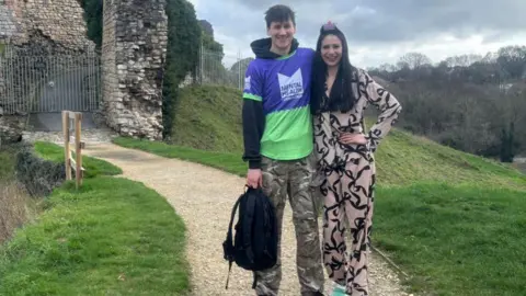 Supplied A young man wearing a black hoodie and camouflaged trousers holding a black backpack and wearing a purple and green top which says 'Mental Health Foundation' on it. He has short brown hair and is smiling with his arm around a woman next to him. The woman has long black hair and is smiling and wearing a two-piece shirt and trousers with black ribbon on it. They are stood on the top of a hill next to some brickwork.