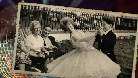 Black and white image from 1963 taken in the Isle of Man of junior dance champions June Batty in a white ballroom dress and John Halliday in a black tuxedo resting on a silver glitterball. They are doing an old-time tango pose while parents sitting on deck chairs look on smiling and Eva holding their winning trophies. 