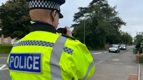 BBC A police officers wears a high vis vest and stands on the pavement taking pictures of speeding cars