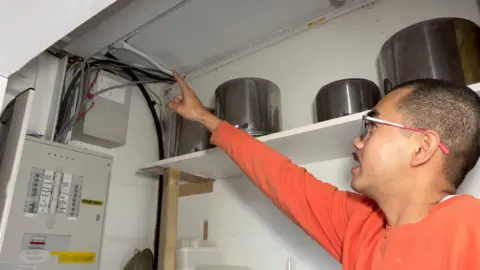 Theo is wearing an orange top and points toward a cluster of cables and wiring near a ceiling, beside metal containers and an electrical panel mounted on the wall.