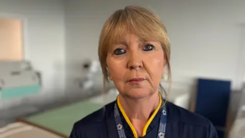 A woman wearing her work uniform stood in a hospital care room. A chair and a hospital bed is visible behind her with medical equipment.