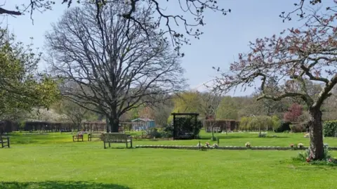 Isle of Wight Council Isle of Wight Crematorium memorial gardens. It has several trees, an expanse of green grass and a number of wooden benches.