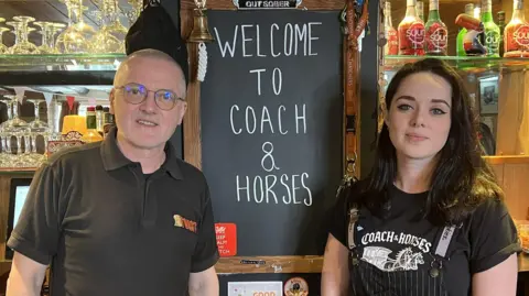 A man with grey hair and glasses and a younger woman with black hair, stripy dungarees and tattoos are standing either side of a black chalkboard which reads 'Welcome to the Coach and Horses.' They both work at the pub and are wearing all black. They are standing behind a bar.