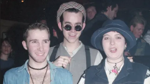 Stuart Linden Rhodes Three people look at the camera. The person on the left wears a light denim, open-neck shirt and necklace. The person in the middle is wearing a hat, sunglasses and grey top. The person on the right is wearing a sleeveless denim jacket and matching hat.