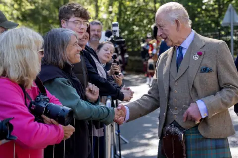 PA Media King Charles III greets members of the public gathered behind crowd barriers during a visit, shaking hands with one person while others look on and take photographs. The King is wearing a tweed jacket, waistcoat and tartan kilt. Several spectators hold cameras and smartphones as they line the roadside beneath trees in bright sunshine.