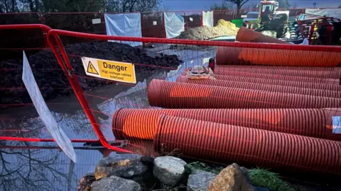 Stephen Goodridge A photo taken at nighttime of the roadworks. There is a red metal tall fence, which has a white and yellow sign which reads 'Danger, high voltage cables overhead'. There is water covering the road and to the right of the image a pile of large red metal tubes. There is a digger in the background and piles of rubble to the left of the image.