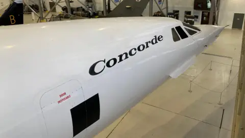 Dave Webster/BBC The front of a Concorde plane is in the image. It is painted white and has small windows at the front, which are on the right of the image, and a sign which says Concorde in black writing. 