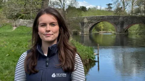 A woman with long dark hair and a blue top with blue and white striped sleeves standing in front of a river with a stone bridge with a number of arches going over it