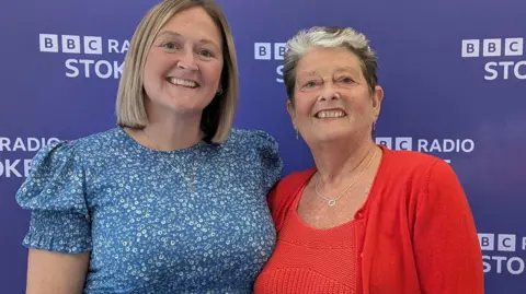 BBC Two women standing together and smiling. The woman on the left has blonde bob hair style and the woman on the right is wearing a red top and red cardigan. She has short hair which is black with a grey fringe. 
