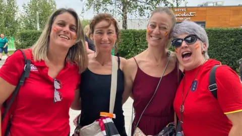 BBC Four women looking into the camera, smiling. Ones on the left and right of the group are wearing red t-shirts, either side of a woman wearing a black vest top and one with a burgundy vest top. They all have small Spanish flags in face paint on their cheeks.