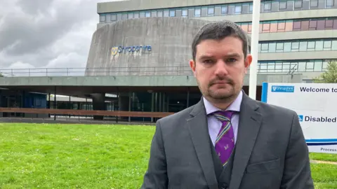 A man wearing a dark grey suit and purple shirt and tie stands in front of a council building. In the middle distance is a building with a concrete cylinder on top with a sign that says Shropshire. Behind that is another building made up of concrete and glass