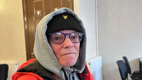 Kate Bradbrook/BBC A man wearing a thick grey hoodie with the hood covering his head and a red puffer coat on top. He has black frame glasses and is looking at the camera. There are black chairs and a door behind him. 