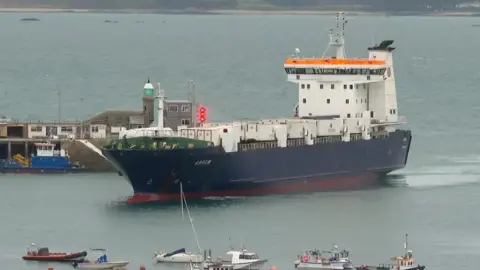 The MV Arrow, a navy blue and white freight vessel. It is pictured sailing into harbour, with a number of smaller boats visible in the foreground. On the side of the vessel it says 'Arrow' in white, capital letters.