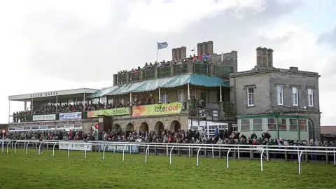 The main stand at Kelso racecourse with big crowds outside and a Scotland flag flying