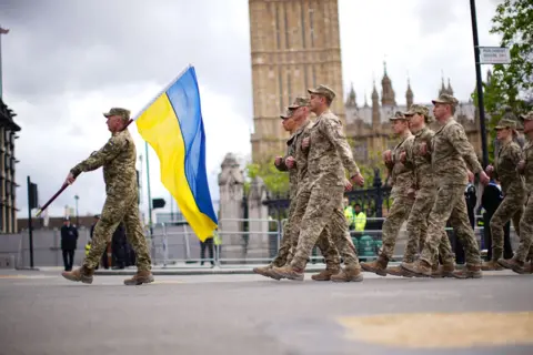 PA Media Military personnel wearing camouflage uniforms walk through Parliament Square while carrying a blue and yellow Ukrainian flag with Big Ben in the background.