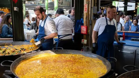 Getty Images Large pans of street food paella at Borough Market. A giant cooking vessel is filled with yellow rice and liquid and men wearing aprons and gloves stir and serve it