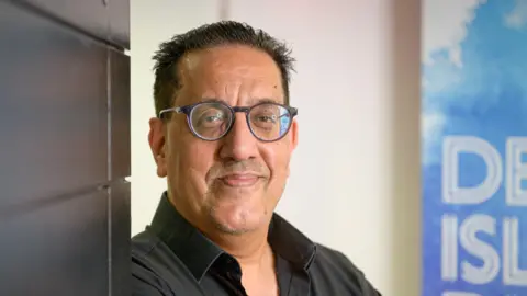 Nazir Afzal, who wears dark glasses and a black shirt, smiles as he leans against a dark wall in front of a blue banner for BBC's Desert Island Discs.