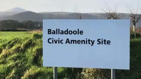 A white sign in front of fields. Black bold writing reads: Balladoole Civic Amenity Site.