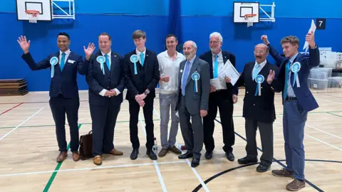 A group of men in suits stand in a sports hall. Some of the men have their arms raised in celebration.