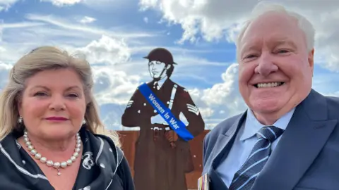 Sarah-May Buccieri/ BBC David Hourigan and his sister, Elizabeth, standing in front of a metal silhouette statue of a female soldier wearing a blue sash that reads "Women in War." Elizabeth stands on the left wearing a black blouse with a patterned collar, with a pearl necklace and pearl earrings. She has long blonde hair. David is standing on the right wearing a navy suit with a blue shirt and a dark and light blue striped tie. He is smiling at the camera and has short white hair. The sky is blue with light clouds. 