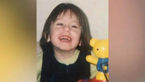 Supplied A smiling child playing with a yellow bear toy in a grainy family photo
