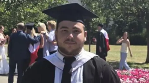 Oliver, who has dark hair and short facial hair, is wearing graduation robes and a cap, standing beside a flower bed in a park, with other graduates in the background.