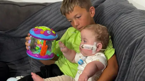 Alfie is sitting on a grey sofa with baby Teddy on his knee. Alfie is wearing a bright lime green T-shirt. His left arm is around his brother's back and he's holding a toy for Teddy to play with in the other hand. The toy is a multi-coloured fish tank which Teddy is reaching out to grab. His feeding tube is taped to his left cheek, and you can see his tracheostomy tube jutting about three inches out from under his chin. It is clear with a blue cap on the end. Teddy is wearing cappuccino coloured leggings and a cream bodysuit with animal patterns.