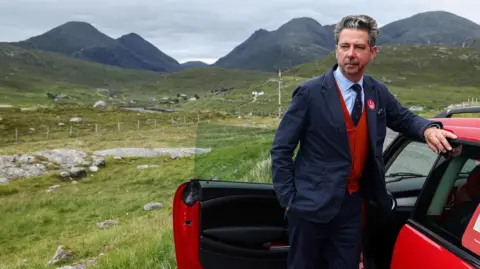 Getty Images Torcuil Crichton is standing outside with hills in the background. He is leaning on a red car. He has his black and grey hair combed back and has a neatly trimmed beard. He is wearing a dark blue suit over a red waistcoat and a blue shirt and a dark tie.