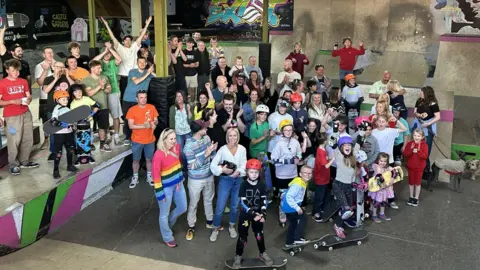 Exist Skatepark A group of people with skateboards and equipment look up at a camera standing on various ramps.