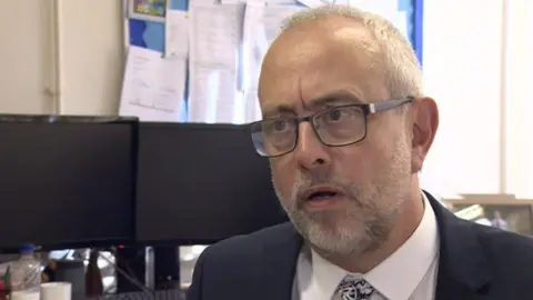 Dave Lee-Allen is pictured mid-interview talking in an office setting. He has thin grey hair as well as a grey beard. He wears a black suit with a white shirt and tie.