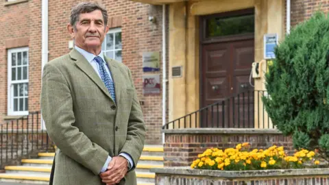 PCC Philip Wilkinson wearing a khaki coloured blazer and blue shirt with his hands together in front of him. He is smiling at the camera and standing in front of the Wiltshire PCC office.