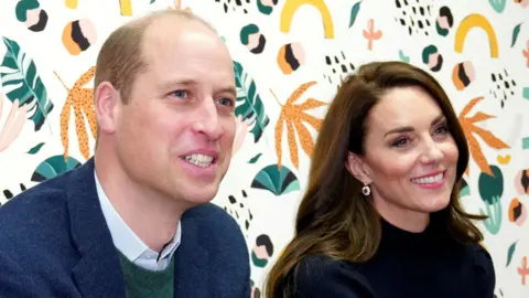 Prince William wearing a blue shirt with a green jumper and blue jacket. Next to him is Kate, the Princess of Wales who is wearing a dark top, she has long brown hair and is wearing a pair of dangly earrings. They are both smiling but looking away from the camera.