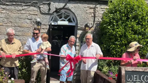 A group of people are pictured standing behind a red ribbon that is wrapped around a pub.