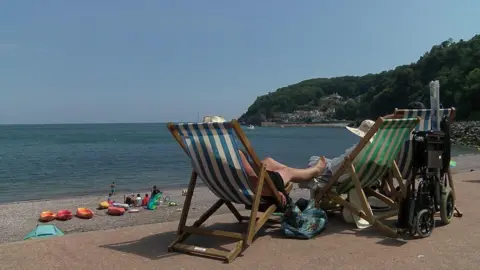 Two people relaxing in deck chairs overlooking a sandy beach. One person is resting their legs on the other person's chair. Calm sea water is lapping on the sandy shoreline. It appears to be a warm day. Four kayaks are visible on the beach in the background. 