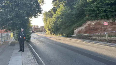 Alice Cunningham/BBC An officer standing by the police cordon on Burrell Road in Ipswich